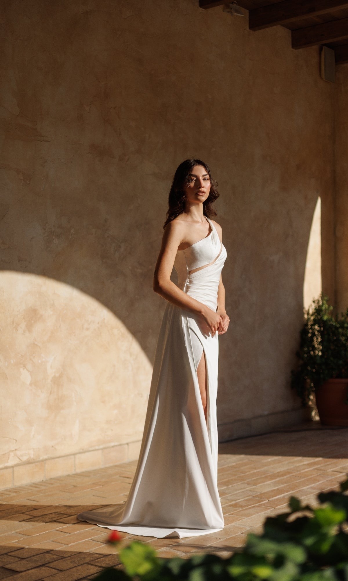 A woman stands in a sunlit corridor wearing the Tina Holly TT001W One-Shoulder White Prom Gown, featuring an elegant off-white hue and a daring thigh-high slit.